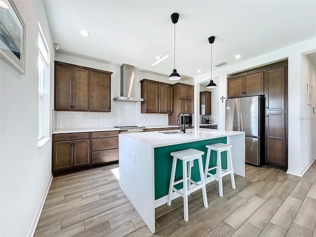 kitchen with pendant lighting, wall chimney exhaust hood, sink, stainless steel fridge, and a kitchen island with sink