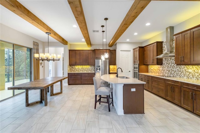 kitchen featuring a center island with sink, hanging light fixtures, wall chimney exhaust hood, appliances with stainless steel finishes, and beam ceiling