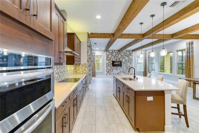 kitchen with a kitchen island with sink, sink, beam ceiling, a stone fireplace, and hanging light fixtures