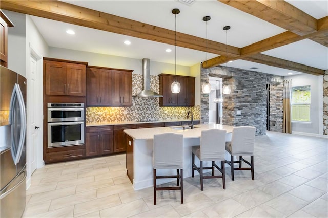 kitchen featuring beam ceiling, wall chimney exhaust hood, stainless steel appliances, hanging light fixtures, and a kitchen island with sink