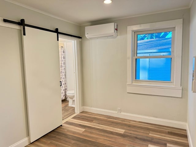 unfurnished bedroom with ensuite bath, ornamental molding, a wall unit AC, a barn door, and hardwood / wood-style flooring