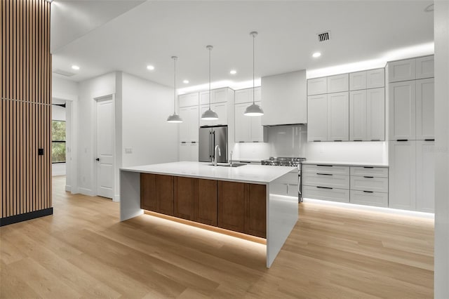 kitchen featuring high end refrigerator, a center island with sink, sink, hanging light fixtures, and light wood-type flooring