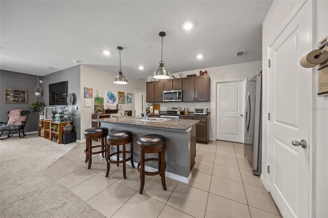 kitchen with dark brown cabinetry, stainless steel appliances, light tile patterned floors, decorative light fixtures, and an island with sink