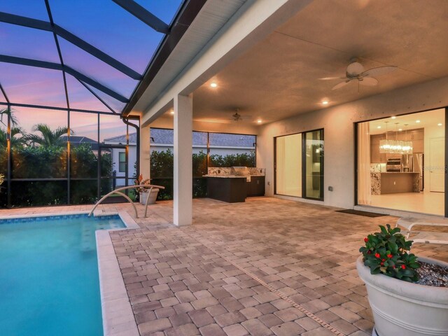 pool at dusk with glass enclosure, an outdoor pool, a ceiling fan, an outdoor kitchen, and a patio