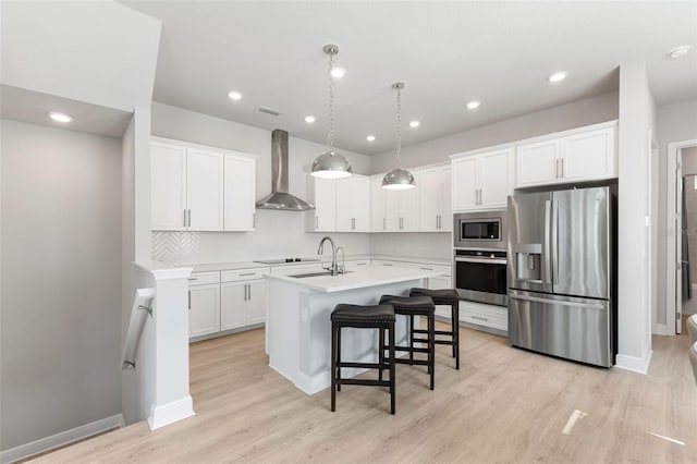 kitchen featuring wall chimney exhaust hood, stainless steel appliances, pendant lighting, a center island with sink, and white cabinets