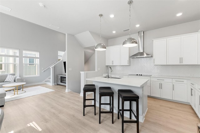 kitchen with wall chimney exhaust hood, sink, a center island with sink, white cabinetry, and hanging light fixtures