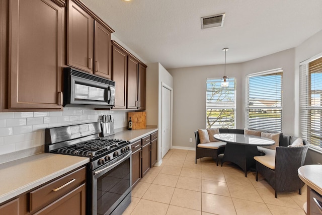 kitchen featuring decorative backsplash, stainless steel gas range oven, dark brown cabinets, light tile patterned floors, and decorative light fixtures