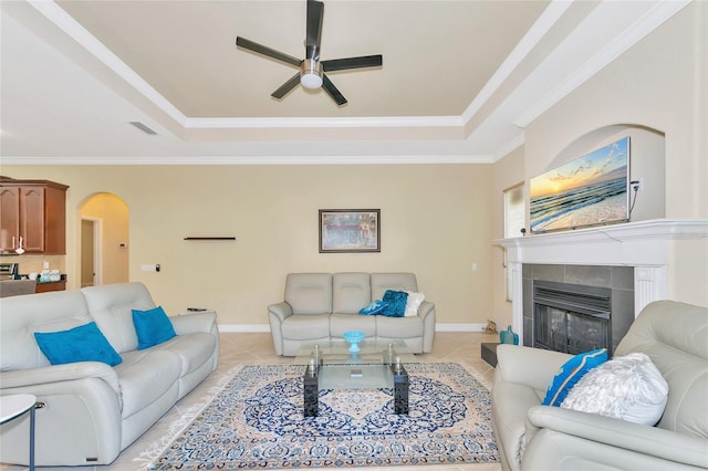 tiled living room featuring a tray ceiling, a tiled fireplace, ceiling fan, and crown molding