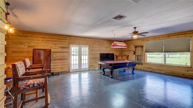 recreation room with ceiling fan, a textured ceiling, wooden walls, and french doors