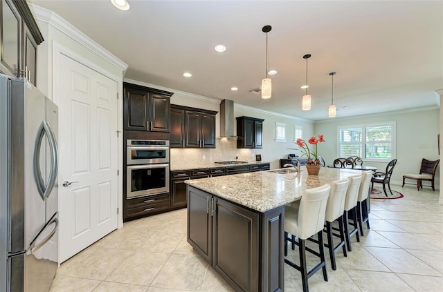 kitchen with a center island with sink, appliances with stainless steel finishes, pendant lighting, ornamental molding, and wall chimney exhaust hood