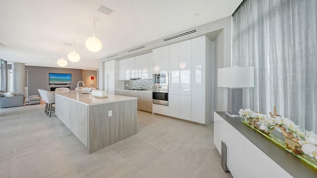 kitchen featuring a center island with sink, white cabinets, decorative backsplash, light tile patterned floors, and decorative light fixtures