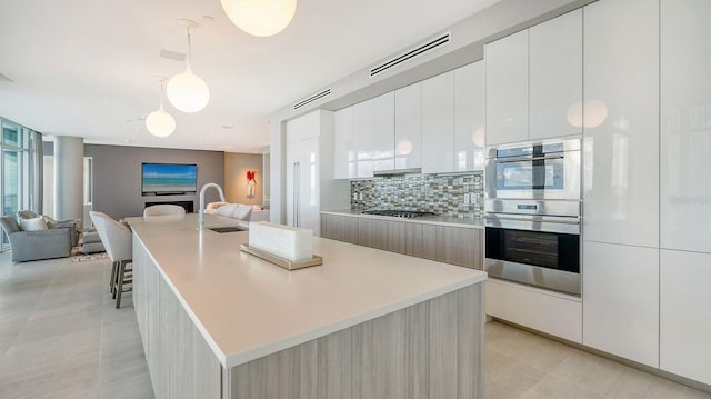 kitchen with tasteful backsplash, a kitchen island with sink, white cabinets, and decorative light fixtures