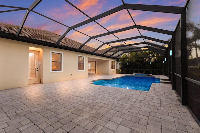 pool at dusk featuring glass enclosure, a patio area, and an in ground hot tub