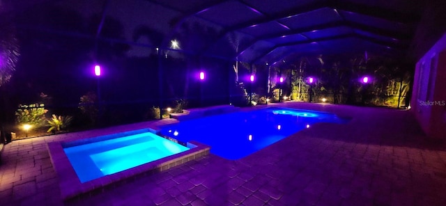 pool at night with an in ground hot tub, glass enclosure, and a patio area