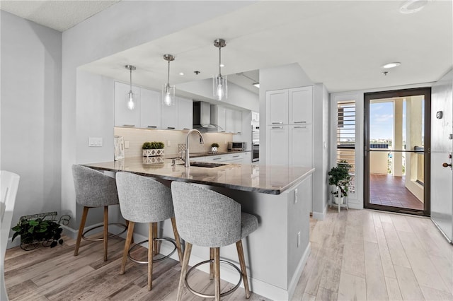 kitchen featuring sink, wall chimney range hood, kitchen peninsula, pendant lighting, and white cabinets