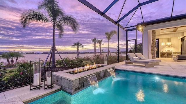 pool at dusk featuring pool water feature, a water view, a fire pit, glass enclosure, and a patio area