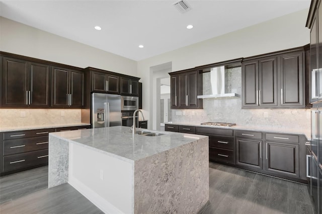 kitchen with wall chimney exhaust hood, an island with sink, dark wood-type flooring, and appliances with stainless steel finishes