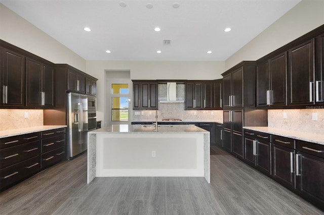 kitchen featuring wall chimney range hood, stainless steel refrigerator with ice dispenser, wood-type flooring, a center island with sink, and dark brown cabinets