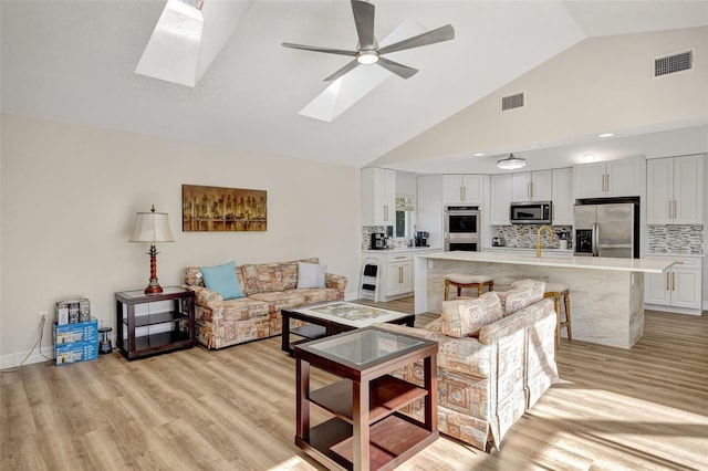 living room with a skylight, ceiling fan, sink, high vaulted ceiling, and light hardwood / wood-style flooring