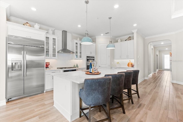 kitchen with pendant lighting, wall chimney range hood, light wood-type flooring, an island with sink, and appliances with stainless steel finishes