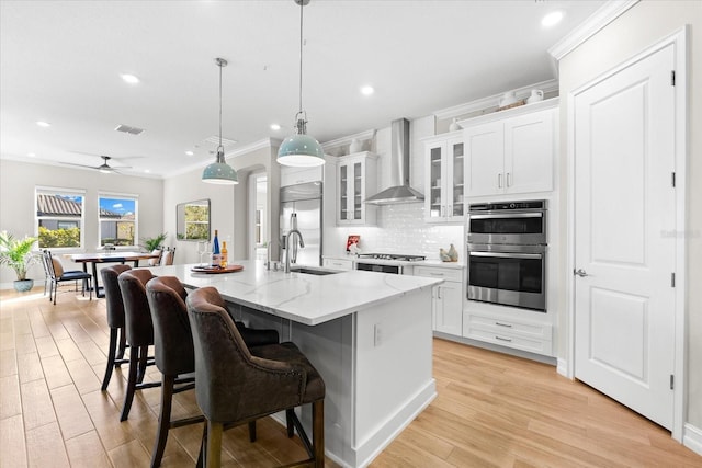 kitchen with a kitchen island with sink, white cabinets, wall chimney exhaust hood, ceiling fan, and stainless steel appliances