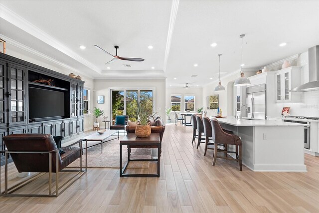 living room featuring a raised ceiling, ceiling fan, light wood-style flooring, ornamental molding, and recessed lighting