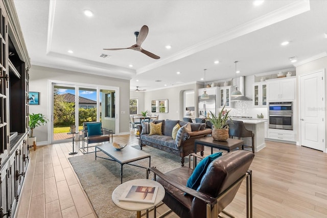 living room featuring light wood finished floors, a raised ceiling, visible vents, ornamental molding, and a ceiling fan