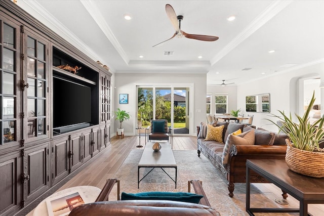 living room with a tray ceiling, visible vents, light wood-style flooring, ornamental molding, and a ceiling fan