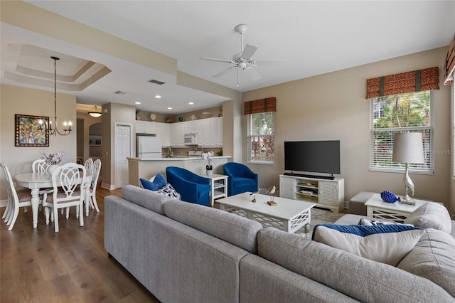 living room with ceiling fan with notable chandelier, a raised ceiling, and dark wood-type flooring