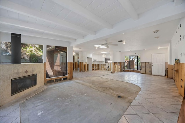 unfurnished living room with a tiled fireplace, ceiling fan, wooden walls, and beamed ceiling