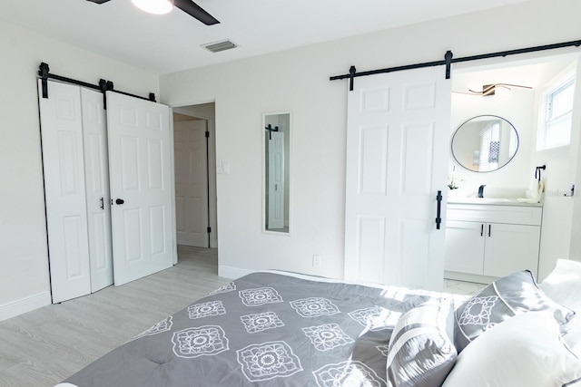 bedroom featuring connected bathroom, a barn door, ceiling fan, and light hardwood / wood-style flooring