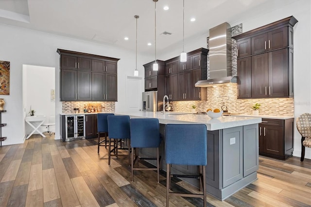 kitchen with a center island with sink, wall chimney range hood, sink, hanging light fixtures, and wood-type flooring