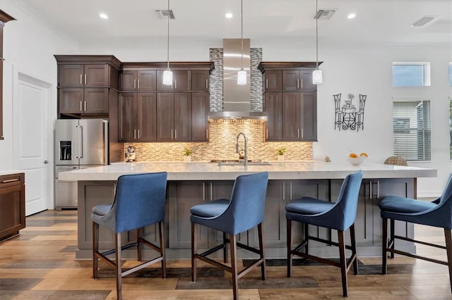 kitchen featuring a center island with sink, hanging light fixtures, wall chimney exhaust hood, high quality fridge, and wood-type flooring