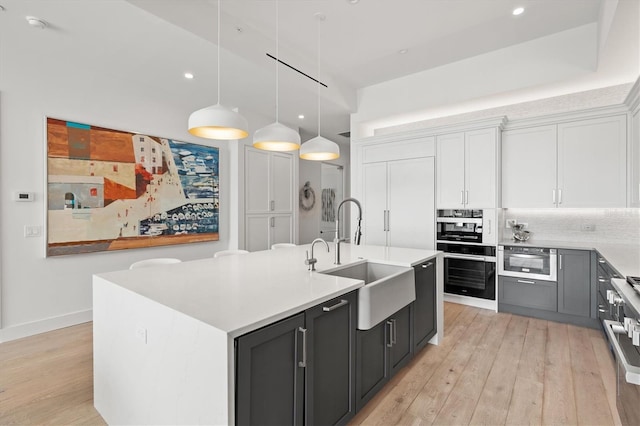 kitchen featuring sink, hanging light fixtures, a kitchen island with sink, white cabinets, and light wood-type flooring