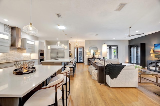 interior space with backsplash, a center island with sink, hanging light fixtures, light hardwood / wood-style flooring, and wall chimney exhaust hood