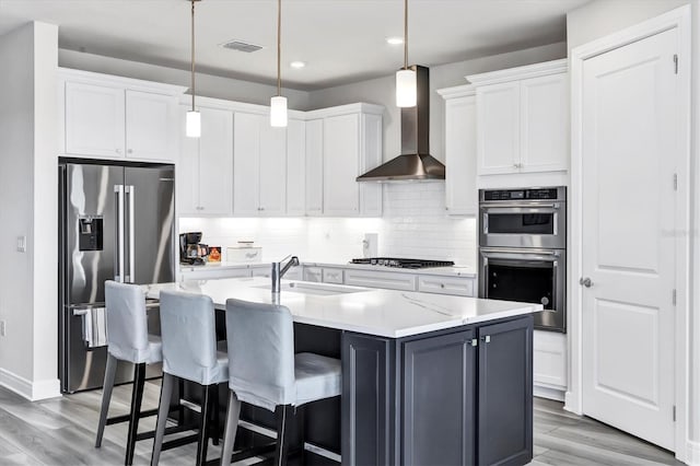 kitchen with white cabinets, appliances with stainless steel finishes, wall chimney range hood, an island with sink, and sink