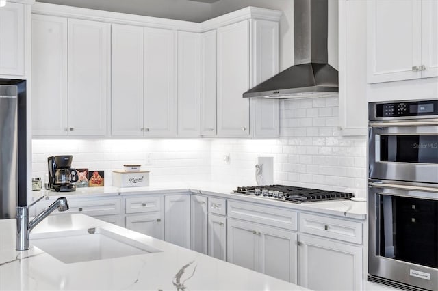 kitchen featuring appliances with stainless steel finishes, wall chimney exhaust hood, white cabinetry, decorative backsplash, and sink