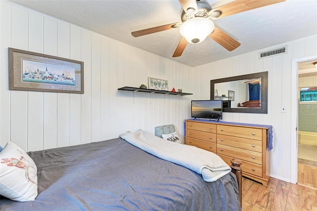 bedroom featuring connected bathroom, light hardwood / wood-style floors, ceiling fan, and wooden walls