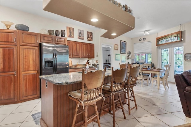 kitchen with a kitchen island with sink, dark stone counters, a kitchen breakfast bar, light tile patterned floors, and stainless steel fridge with ice dispenser