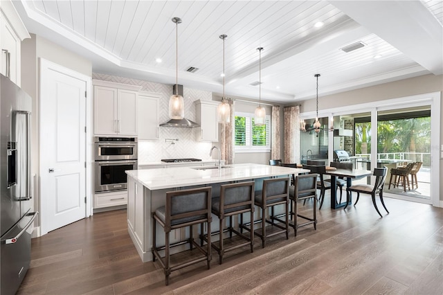 kitchen featuring wall chimney exhaust hood, stainless steel appliances, dark wood-type flooring, a center island with sink, and white cabinets