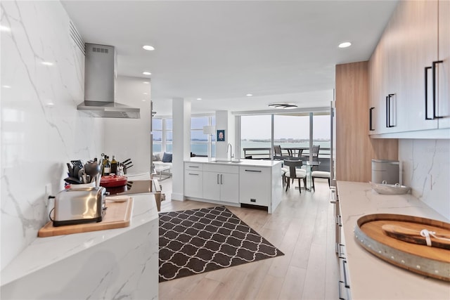 kitchen with light stone counters, wall chimney exhaust hood, sink, light hardwood / wood-style flooring, and white cabinets