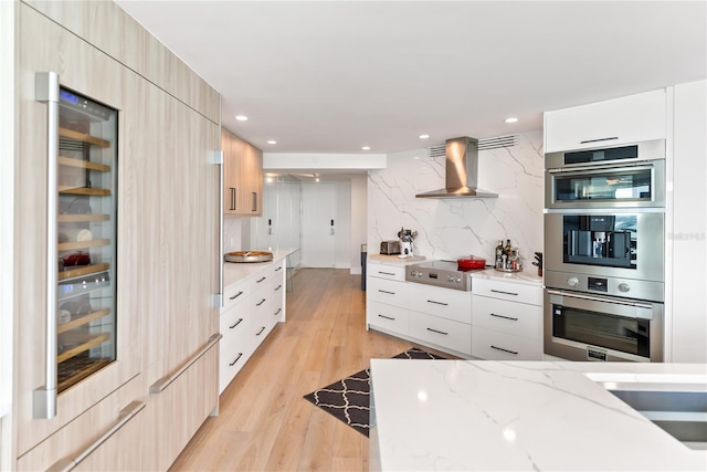 kitchen with wall chimney exhaust hood, stainless steel double oven, cooktop, light hardwood / wood-style flooring, and light brown cabinetry