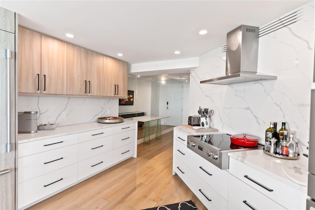 kitchen with wall chimney range hood, tasteful backsplash, light brown cabinetry, stainless steel cooktop, and light wood-type flooring