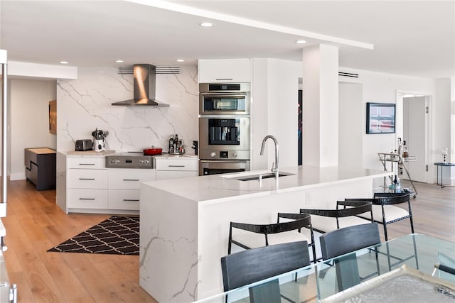 kitchen featuring a kitchen bar, wall chimney exhaust hood, stainless steel appliances, white cabinets, and light hardwood / wood-style floors