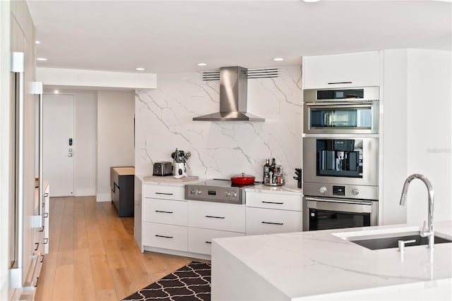 kitchen featuring light stone countertops, wall chimney exhaust hood, stainless steel appliances, sink, and white cabinetry