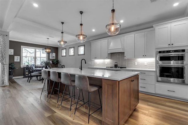 kitchen featuring custom exhaust hood, appliances with stainless steel finishes, hanging light fixtures, a kitchen island with sink, and white cabinets