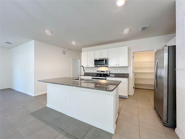 kitchen featuring stainless steel appliances, sink, dark stone countertops, white cabinets, and an island with sink