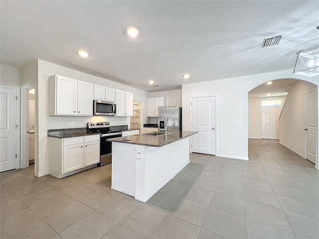 kitchen featuring appliances with stainless steel finishes, a textured ceiling, a center island with sink, dark stone countertops, and white cabinets