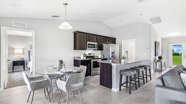 kitchen with dark brown cabinetry, stainless steel appliances, vaulted ceiling, a center island with sink, and stone counters