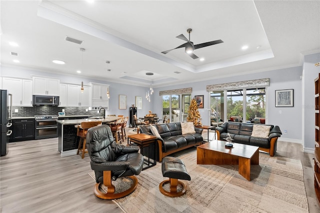 living room with ceiling fan, sink, light hardwood / wood-style floors, a tray ceiling, and ornamental molding